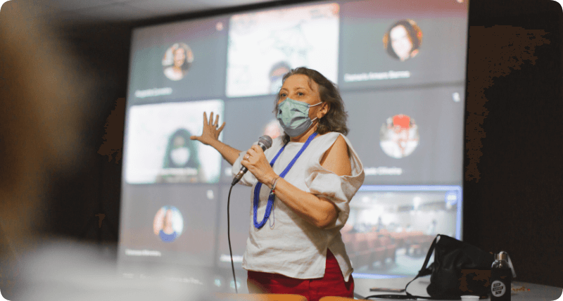 Foto de professora Bete Jaguaribe falando ao microfone. Ela usa máscara.