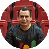 Foto de Adriano Loureiro, gestor do Centro de Formação Olímpica do Ceará. Adriano é um homem branco e usa blusa na cor preta com detalhes amarelo, vermelho e verde. Ele sorri e está sentado em uma arquibancada com cadeiras vermelhas.