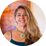 Foto de Charmene Rocha, gestora do Complexo Ambiental Mirante do Caldas. Charmene é uma mulher branca e loira. Ela usa blusa azul marinho de alças finas e um colar verde, com uma flor com detalhes preto e vermelho. Ela sorri e olha para a foto.