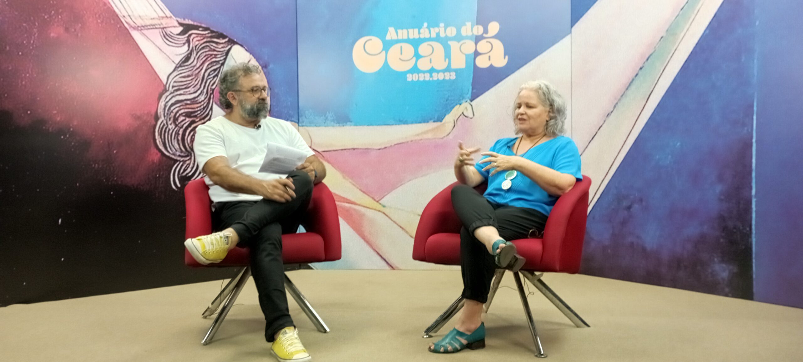 Foto de entrevista em que aparece a presidenta do IDM, Rachel Gadelha. O jornalista que a entrevista é Jocélio Leal. Ambos estão sentados em cadeiras vermelhas e ao fundo lê-se: Anuário do Ceará 2022-2023.