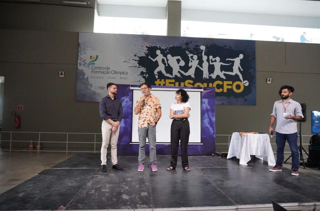 Quatro pessoas em um palco. Ao centro, de camisa estampada branca e laranja e calça jeans, Lenildo Gomes, diretor do IDM, fala ao microfone.