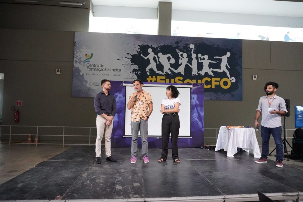 Quatro pessoas em um palco. Ao centro, de camisa estampada branca e laranja e calça jeans, Lenildo Gomes, diretor do IDM, fala ao microfone.