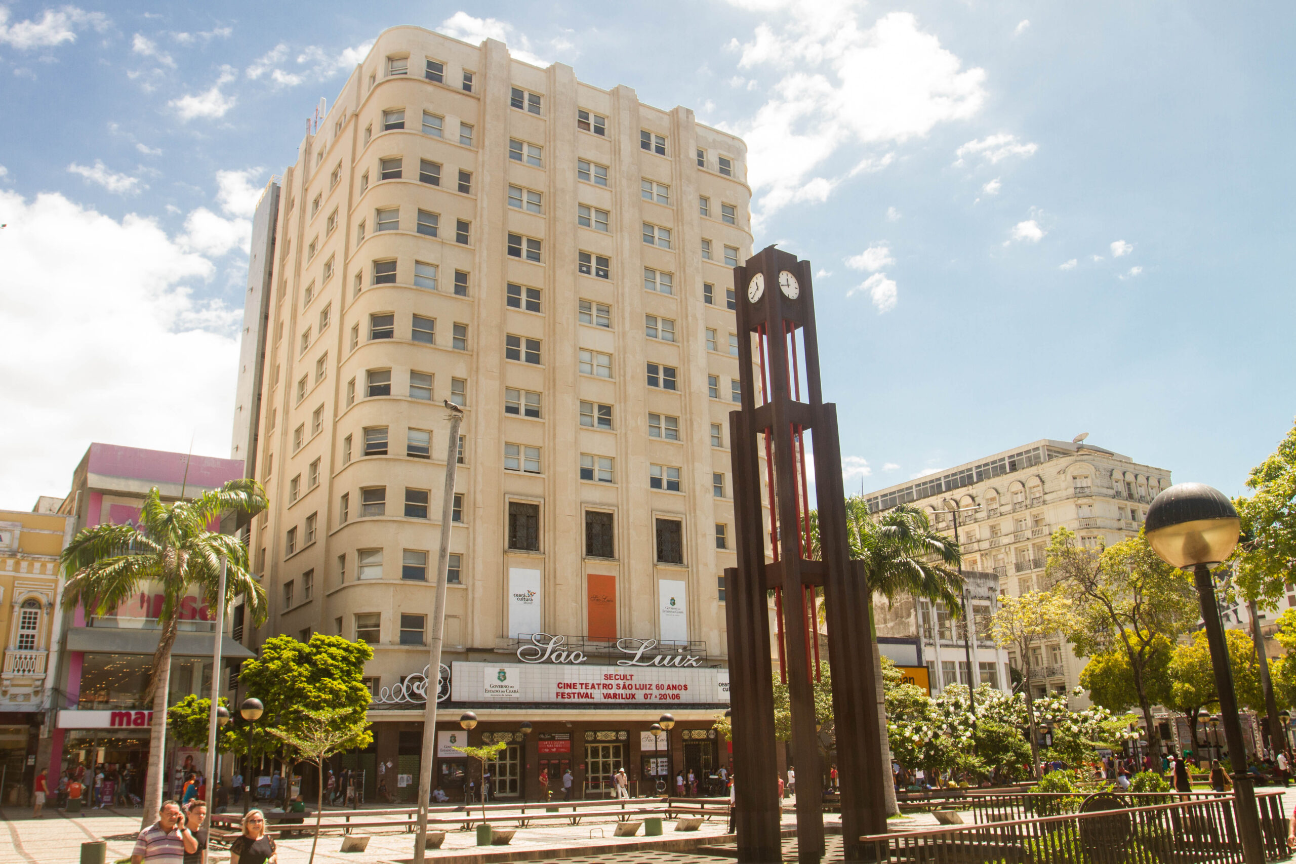 Foto da fachada do Cineteatro São Luiz. É possível ver parte da Praça do Ferreira, onde fica localização do Cineteatro. É possível ver o relógio alto e característico do local.