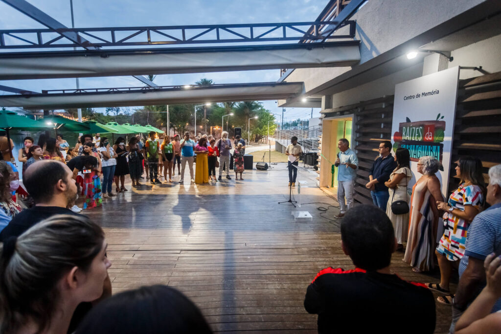Em um espaço aberto, sobre um piso de tábuas de madeira, em frente a uma placa colorida com os dizeres "Centro de Memória Raízes da Sabiaguaba), diversas pessoas, com roupas de cores variadas, estão reunidas.