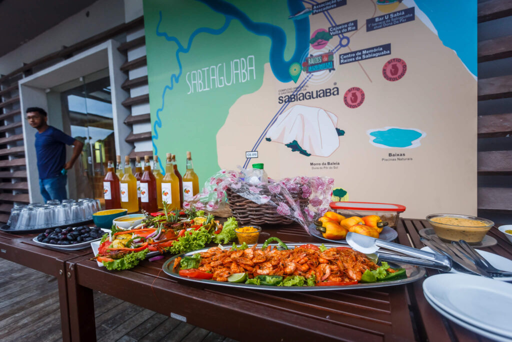 Em frente a um banner com o mapa da Sabiaguaba, em verde, bege, azul e vermelho, uma mesa de madeira marrom-escuro, com alimentos como camarão, caju e frutas. Na extremidade esquerda (ponto de vista do leitor), algumas garrafas com líquidos entre o marrom-escuro e o marrom-claro e rótulos de fundo branco e desenhos laranja.