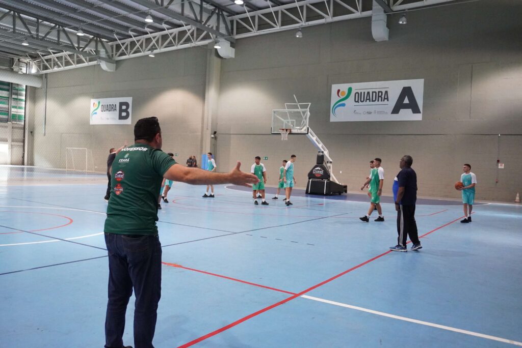 Em uma quadra azul, nove homens jogam basquete vestidos com uniformes  em azul e verde. Mais três homens, vestidos de verde e preto, acompanham de dentro da quadra o jogo.