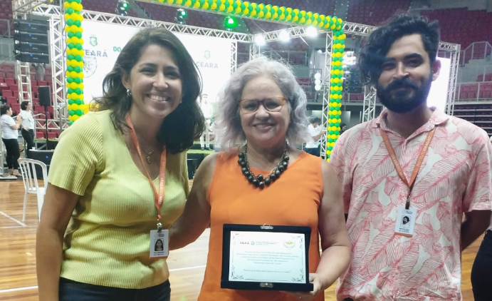 Foto de 3 pessoas que trabalham no IDM segurando uma placa. Duas mulheres, dentre elas a presidenta do IDM, Rachel Gadelha, e um rapaz. Todos estão sorrindo.