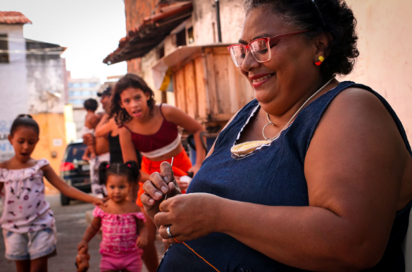 Foto de senhora negra elaborando um bordado. Ela usa óculos e sorri. Ao fundo é possível ver algumas crianças.