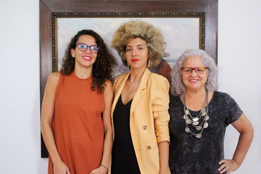 Ao centro, Helena Barbosa, uma mulher preta cis, de cabelos afro pintados de loiro, blazer amarelo e blusa preta. Da perspectiva do leitor, à esquerda, Luísa Cela, uma mulher branca, de cabelos cacheados, óculos e vestido marrom, e, à direita, Rachel Gadelha, uma mulher cis, branca, cabelos brancos, blusa preta, colar e óculos.