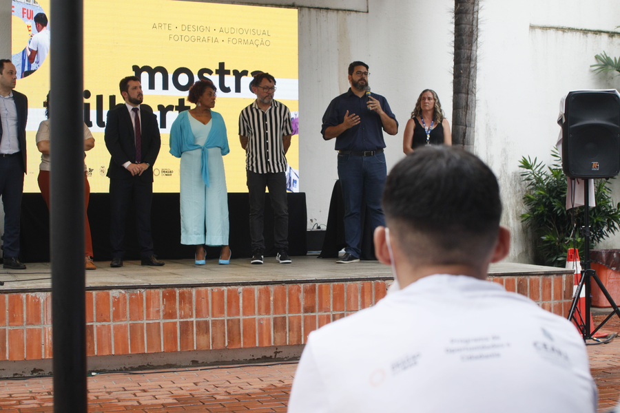 Um homem branco de barba, camisa azul de mangas compridas e óculos fala ao microfone. Ao redor dele homens e mulheres de raças variadas e roupas em diversas cores, escuta. À frente, um jovem de camiseta branca com com logos escuta.