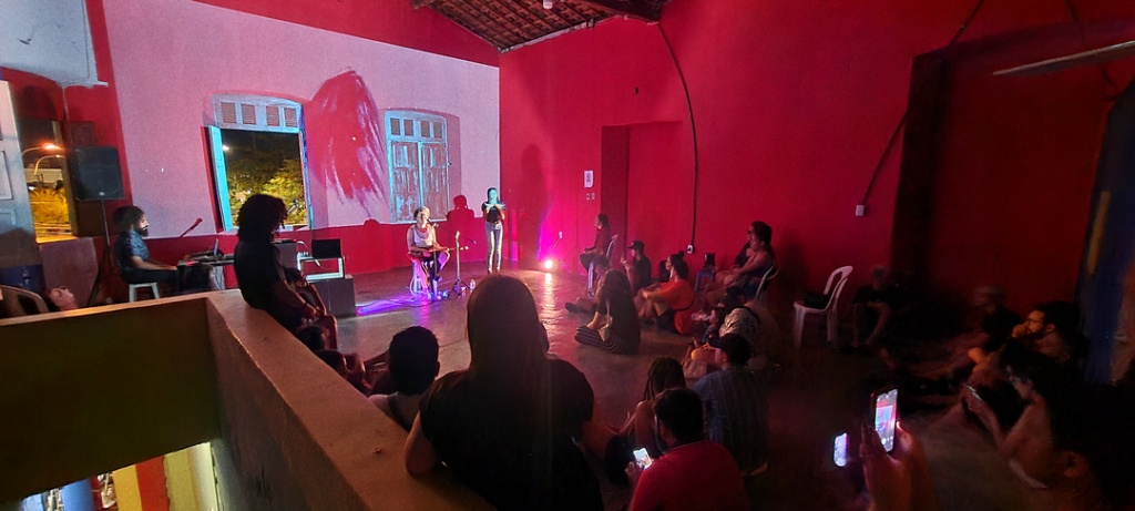 Uma apresentação de música em um ambiente de paredes na cor rosa, com iluminação cênica, com várias pessoas sentadas em semicírculo.