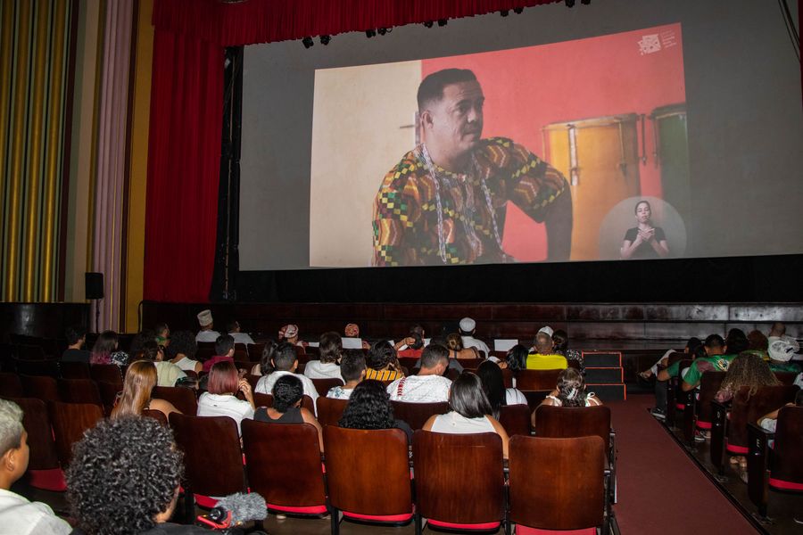 No Cineteatro São Luiz, sentadas em poltronas na cor vinho, pessoas com roupas em cores variadas, algumas trajando vestimentas da Umbanda e Candomblé, assistem na tela o documentário Povos de Terreiro. Na cena, um homem preto, de camisa estampada, fala.