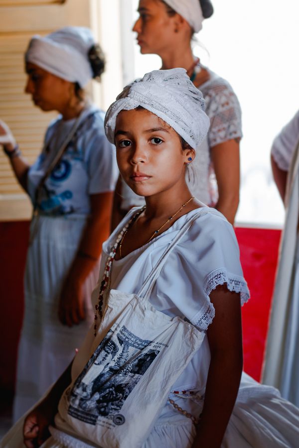 Com algumas mulheres vestidas de branco ao fundo, uma garota preta de pele clara, entre 10 e 12 anos, com turbante e vestido brancos, olha para a câmera