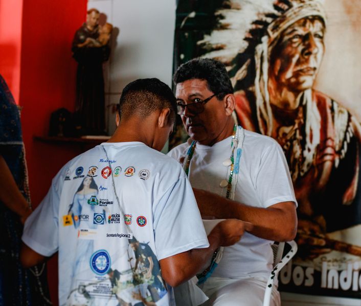 Dois homens pretos de pele clara, vestidos com calças brancas e camisas brancas com logos coloridas nas costas, usando colares de contas, participam de um ritual. Ao fundo, imagens de um santo e de um cacique indígena.