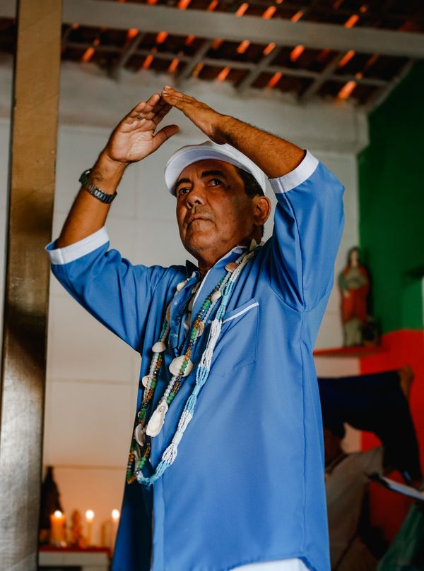 Um homem preto de pele clara, com camisa azul claro de estampa branca e chapéu branco, usando colares de contas, junta as pontas dos dedos acima da cabeça. Ao fundo, desfocados, velas e imagens de santos.