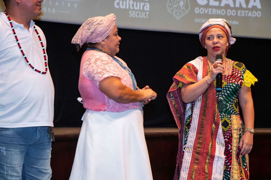 Mãe Janaína, mulher preta, de vestido e turbante coloridos, fala ao microfone. Ao lado dela estão Mãe Sandra, mulher preta de pele clara, com turbante e blusa rosas claro e saia branca, e Pai Giuliano, cujo rosto não aparece todo na foto, vestindo  camisa branca e usando colar de contas colorido