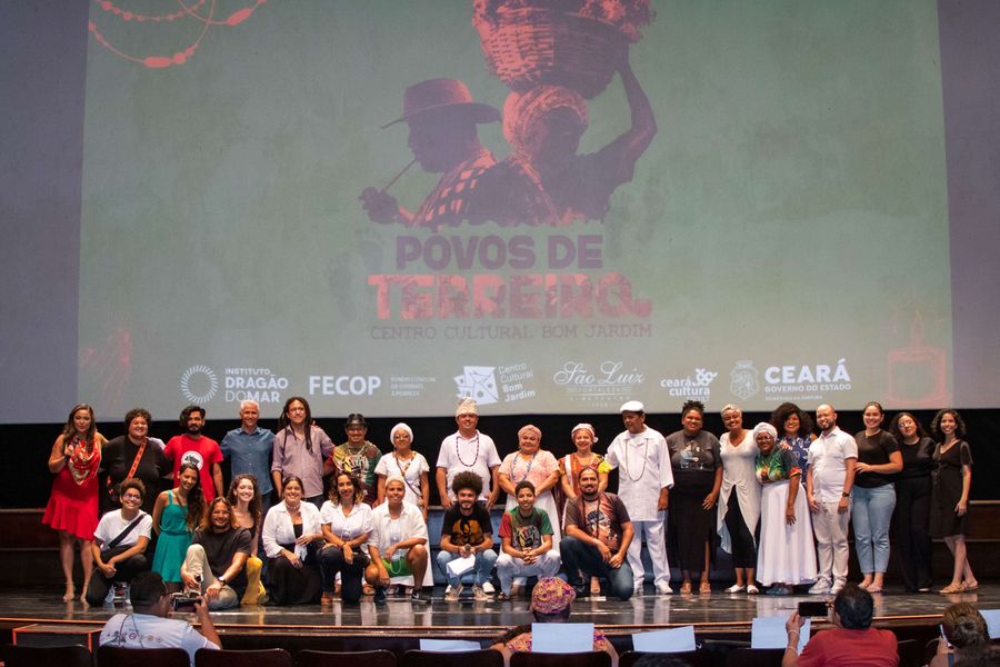 Em frente à tela do Cineteatro São Luiz, sobre o palco, onde se projeta o banner de fundo verde e letras marrons do "Povos de Terreiro", estão as pessoas que participaram da elaboração e difusão do documentário, com roupas de cores variadas.