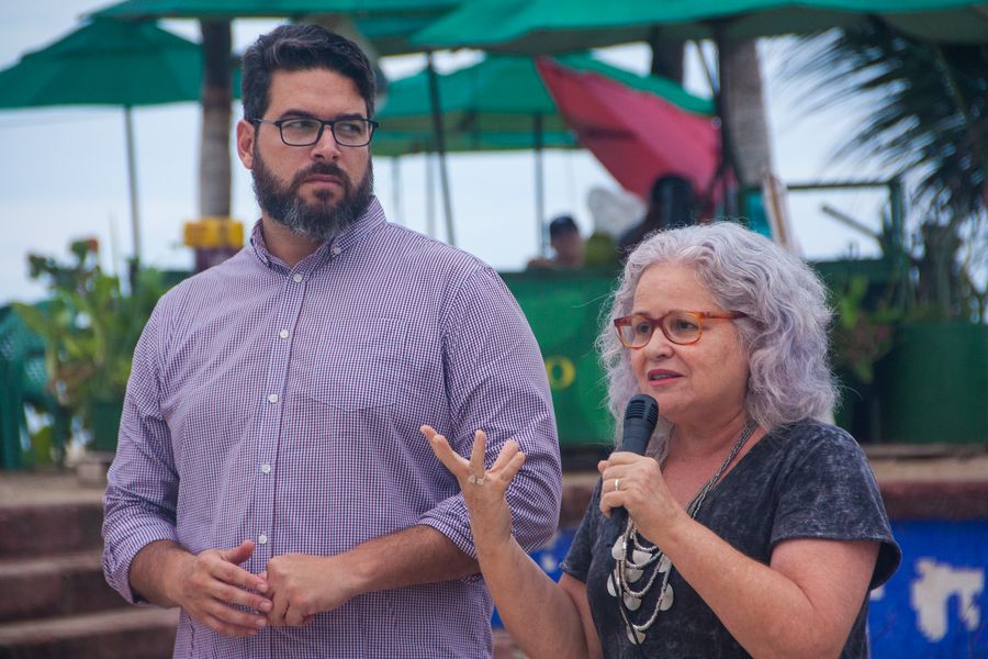 Umamulher de camisa preta, branca e cabelos brancos commicrofone na mão e um homem branco,de óculos, camisa xadrez e barba