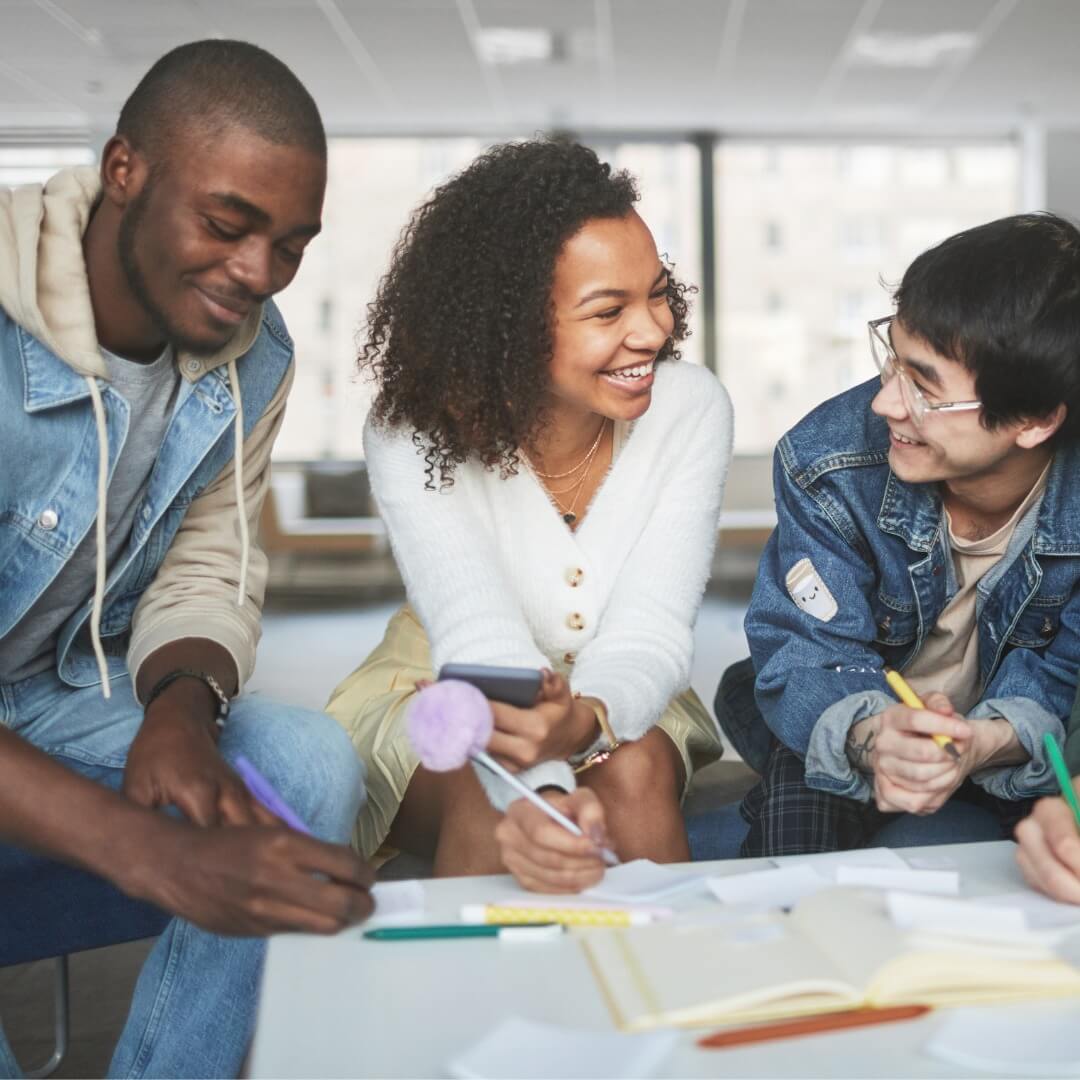 Da esquerda para a direita (perspectiva do leitor): um rapaz preto, sorri, vestindo um moletom bege com capuz, jaqueta e calça jeans; uma jovem preta de camisa branca de manga comprida e saia bege sorri e um rapaz branco de jaqueta jeans, camisa bege e calça xadrez também sorri. Eles se olham entre si, sentados à frente de uma mesa branca com papeis e canetas sobre ela.