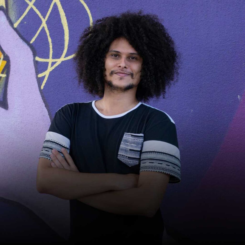 Jovem negro, cabelos afro, camisa preta com detalhes em preto e branco, barba preta, braços cruzados na frente do peito. Foto ilustrativa.