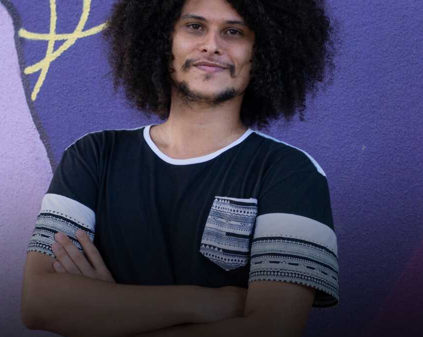 Jovem negro, cabelos afro, camisa preta com detalhes em preto e branco, barba preta, braços cruzados na frente do peito. Foto ilustrativa.