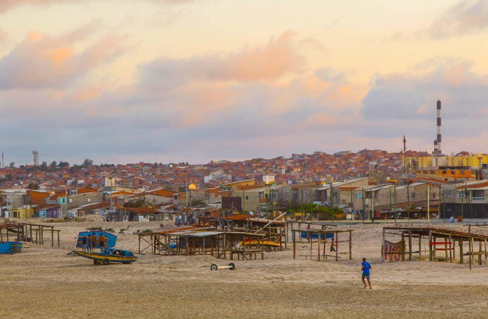 Card com foto de exposição Periferia Presente. É possível ver comunidade na beira da praia. É possível ver barcos, barracas e casas. 