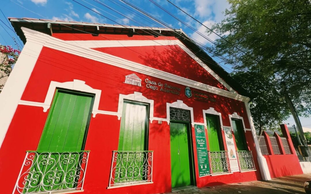 Uma edificação do início do Século XX, com paredes vermelhas, detalhes brancos e portas verdes. A copa verde de uma árvores aparece no canto superior direito.