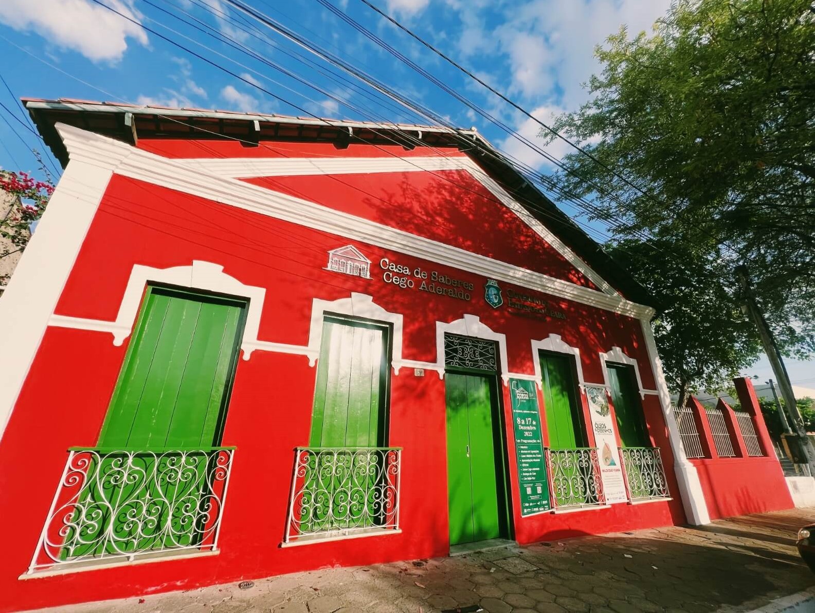 Uma edificação do início do Século XX, com paredes vermelhas, detalhes brancos e portas verdes. A copa verde de uma árvores aparece no canto superior direito.