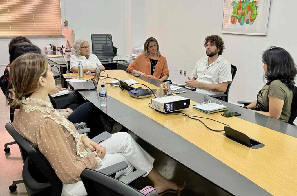 Sete pessoas numa sala em torno de uma mesa em cores cinza e amarela conversam