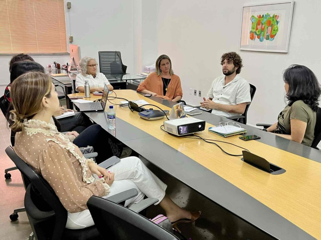 Sete pessoas numa sala em torno de uma mesa em cores cinza e amarela conversam