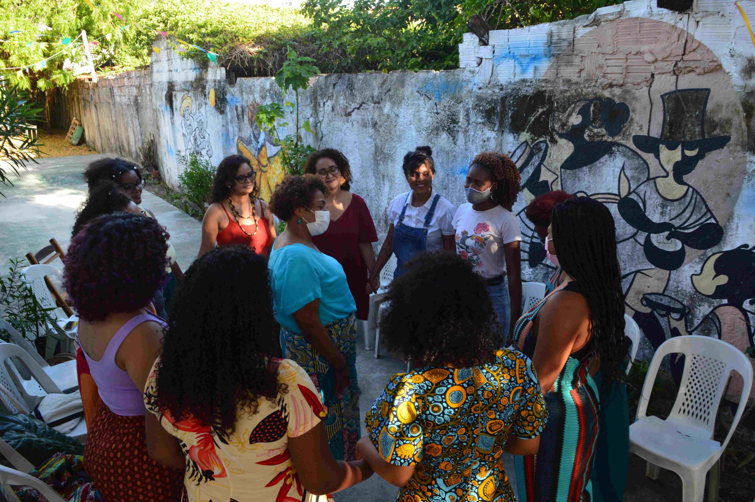 Doze mulheres negras, em roda, de máscaras, cores de cabelos e das roupas variadas, dão as mãos. Ao centro, uma mulher negra, de máscara branca, blusa azul e saia azul estampada. Atrás delas, um muro branco com grafite.