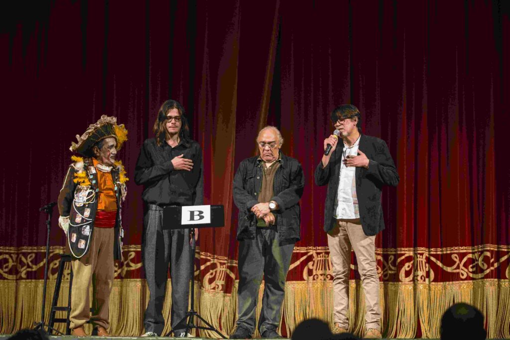 Quatro homens estão no palco do Theatro José de Alencar, com uma cortina vermelha e detalhes dourados ao fundo. Da perspectiva de quem lê, da direita para a esquerda: um homem alto, de cabelo liso castanho, blazer preto, blusa branca e calça bege, um homem calvo, de casaco preto, óculos, as mãos cruzadas na frente da barriga e a expressão triste, um homem branco, de aproximadamente 21 anos, cabelos no ombro, lisos e castanhos, óculos e roupa preta, um homem vestindo fantasia colorida, com o rosto pintado e chapéu artesanal.