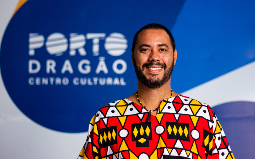 Um homem negro, alto, cabelos e barba, bigode negros, veste uma camisa com estampas coloridas em amarelo e vermelho. Atrás dele, a logo do Porto Dragão (branca sobre um fundo azul).