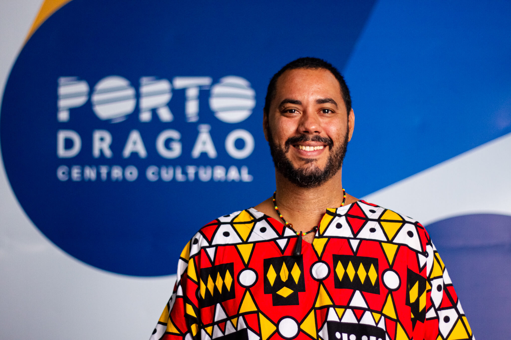 Um homem negro, alto, cabelos e barba, bigode negros, veste uma camisa com estampas coloridas em amarelo e vermelho. Atrás dele, a logo do Porto Dragão (branca sobre um fundo azul).