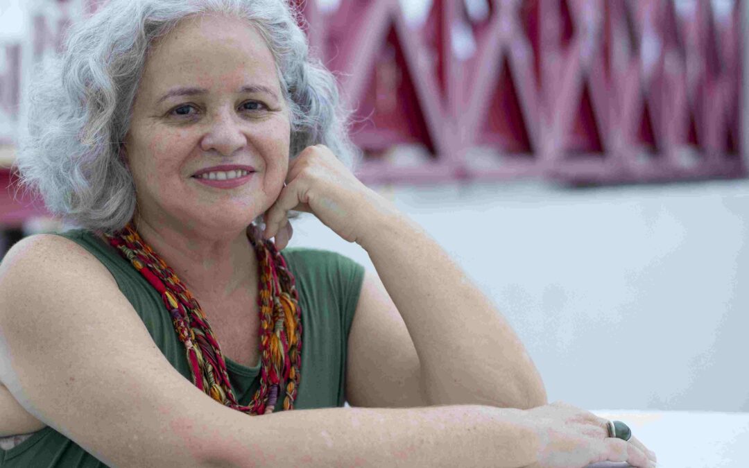 Uma mulher branca, de cabelos brancos curtos, blusa verde, colar colorido, apoia-se num parapeito branco e sorri para a câmera.