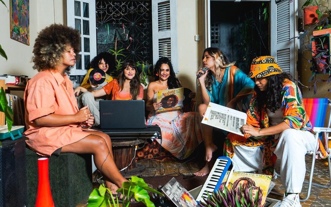 Card com 5 pessoas em uma sala com janela, plantas e bem colorida. As 5 pessoas, integrantes do coletivo de reggae, usam roupas de tons claros.