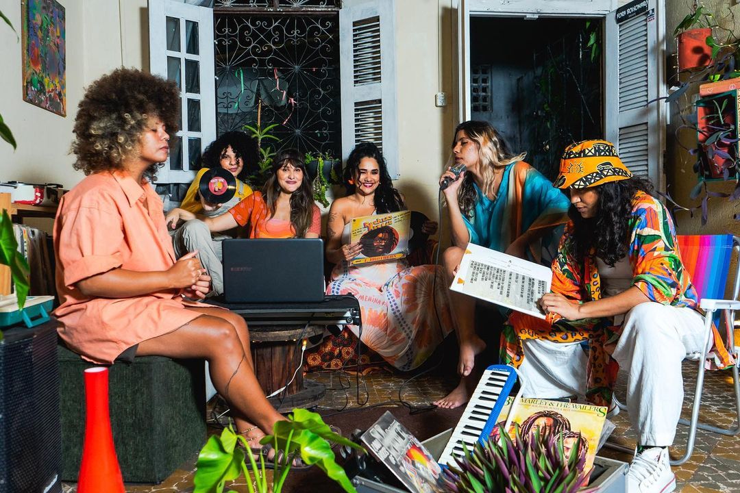 Card com 5 pessoas em uma sala com janela, plantas e bem colorida. As 5 pessoas, integrantes do coletivo de reggae, usam roupas de tons claros.