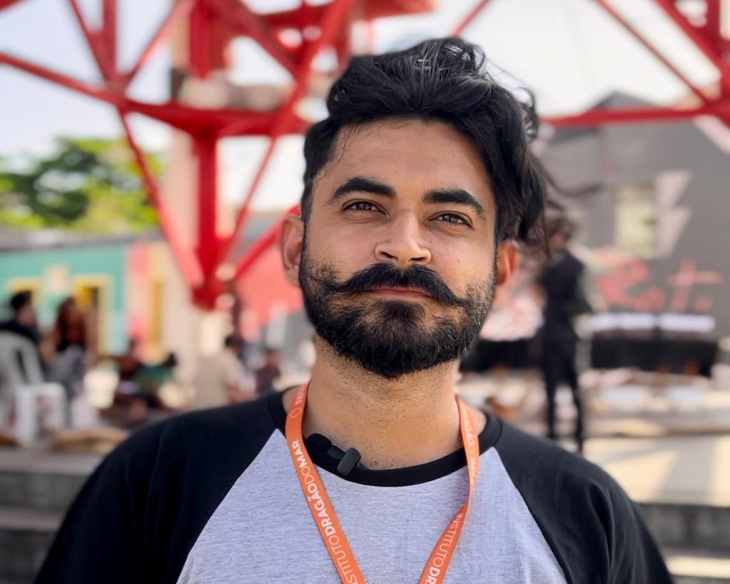 Um homem negro-pardo, barba e bigode pretos, cabelo preto, cordão de crachá laranja, blusa preta com cinza.