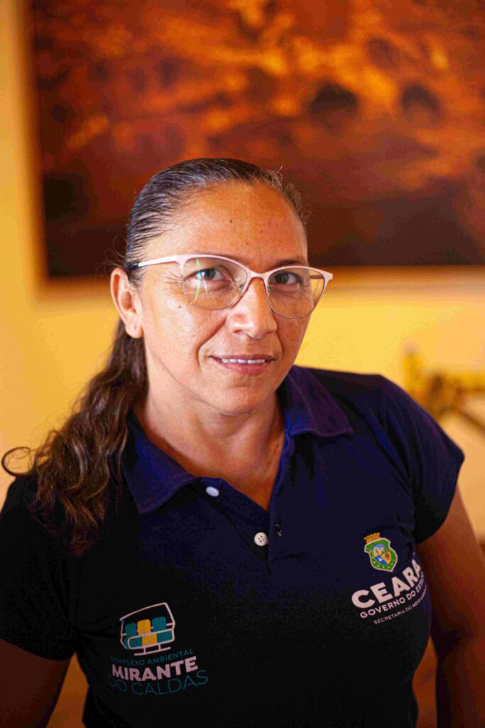 Uma mulher cis branca, de camisa preta, cabelos loiros presos em rabo de cavalo e óculos de grau olha para a câmera.