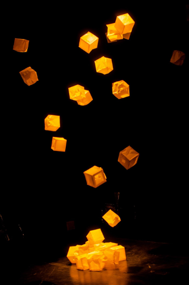 Foto de vários cubos caindo. Uns estão no ar e outros estão no chão. Uma luz laranja ilumina a cena. 