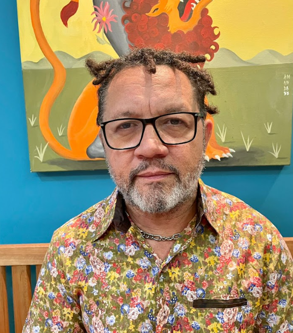Lenildo Gomes usa blusa estampada de flores. Usa óculos, barba e dreads no cabelo. No fundo é possível ver um quadro.