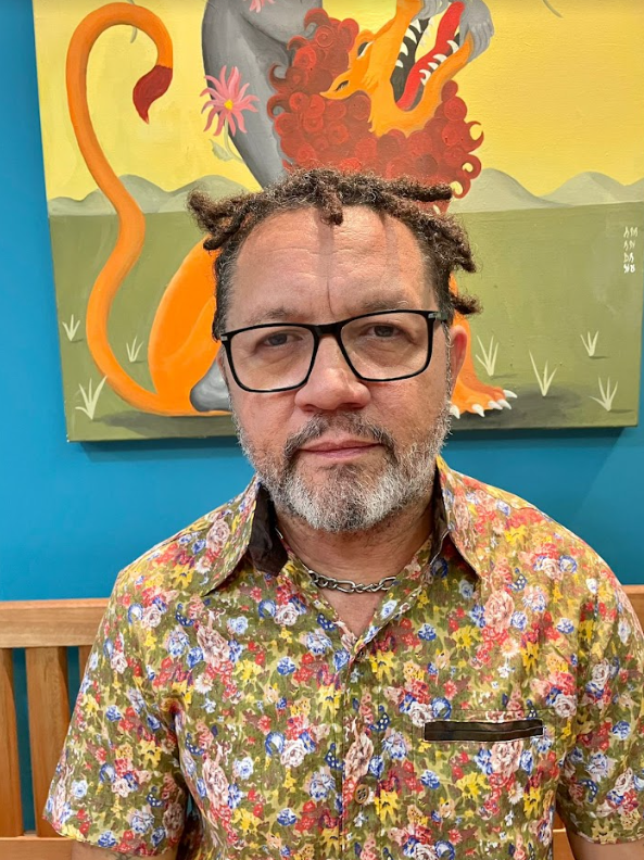 Lenildo Gomes usa blusa estampada de flores. Usa óculos, barba e dreads no cabelo. No fundo é possível ver um quadro.