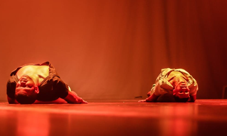 Foto de duas pessoas em espetáculo de dança. Elas estão deitadas no chão. 