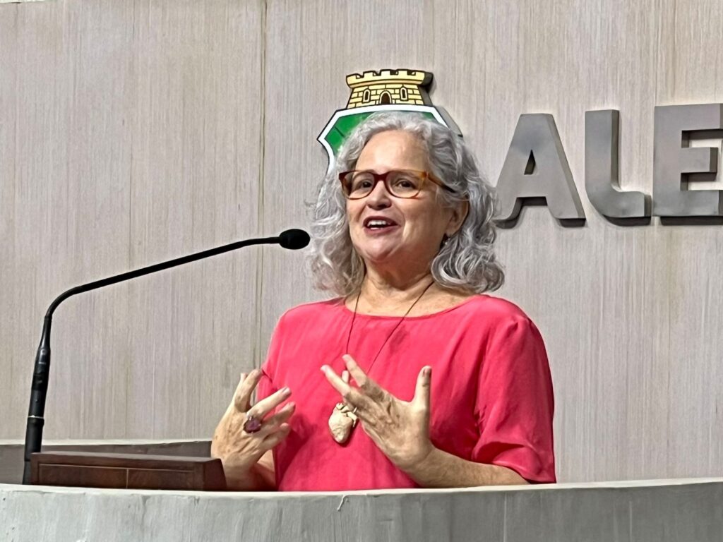 Uma mulher cis, branca, de óculos, cabelos brancos, vestido rosa e colar de coração em marrom, fala gesticulando em um púlpito de mármore.