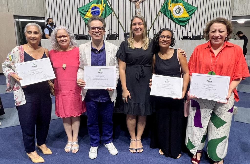Seis pessoas de gêneros, raças e idades diferentes posam para a foto segurando certificados da homenagem recebida na Alece