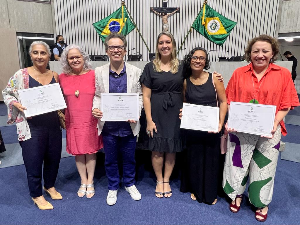 Seis pessoas de gêneros, raças e idades diferentes posam para a foto segurando certificados da homenagem recebida na Alece