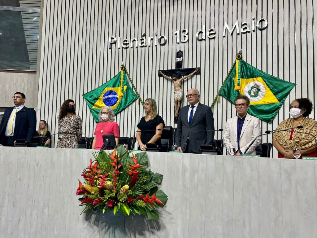 Seis pessoas, de gêneros, raças e idades diferentes, se alinham horizontalmente na mesa composta para Sessão Solene. Elas usam roupas de cores variadas.