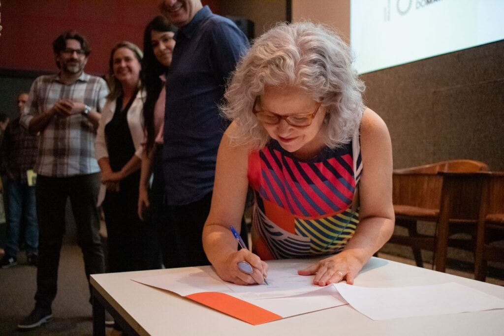 Mulher branca, cis, de óculos, cabelos brancos, blusa estampada, se inclina sobre a mesa e assina, com caneta azul, um documento