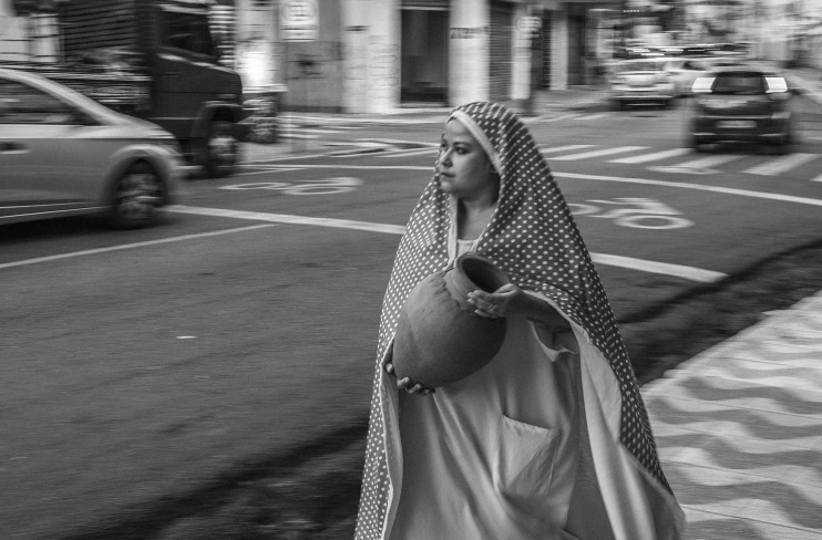 Mulher segurando pote. Parece estar caminhando na rua. Ela usa um longo véu sobre a cabeça.