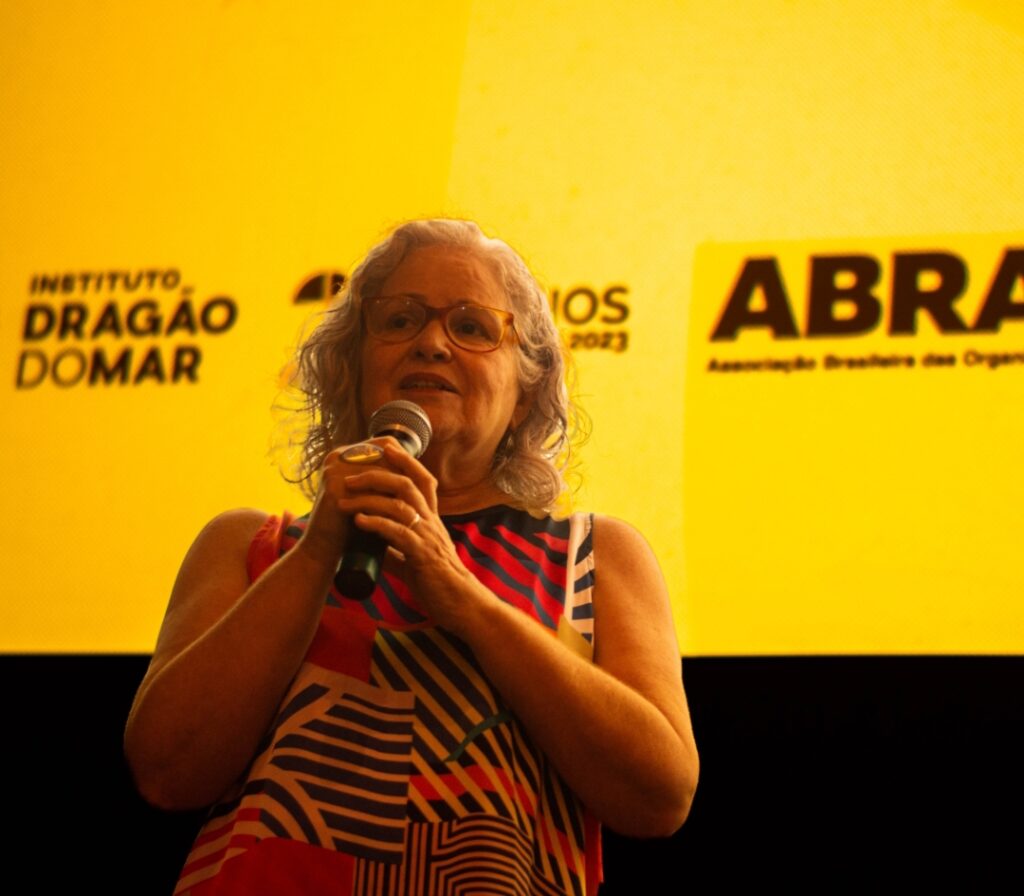 Uma mulher branca, cis, de óculos e camisa colorida, cabelos brancos, fala ao público segurando um microfone. Atrás dela, um fundo amarelo. 